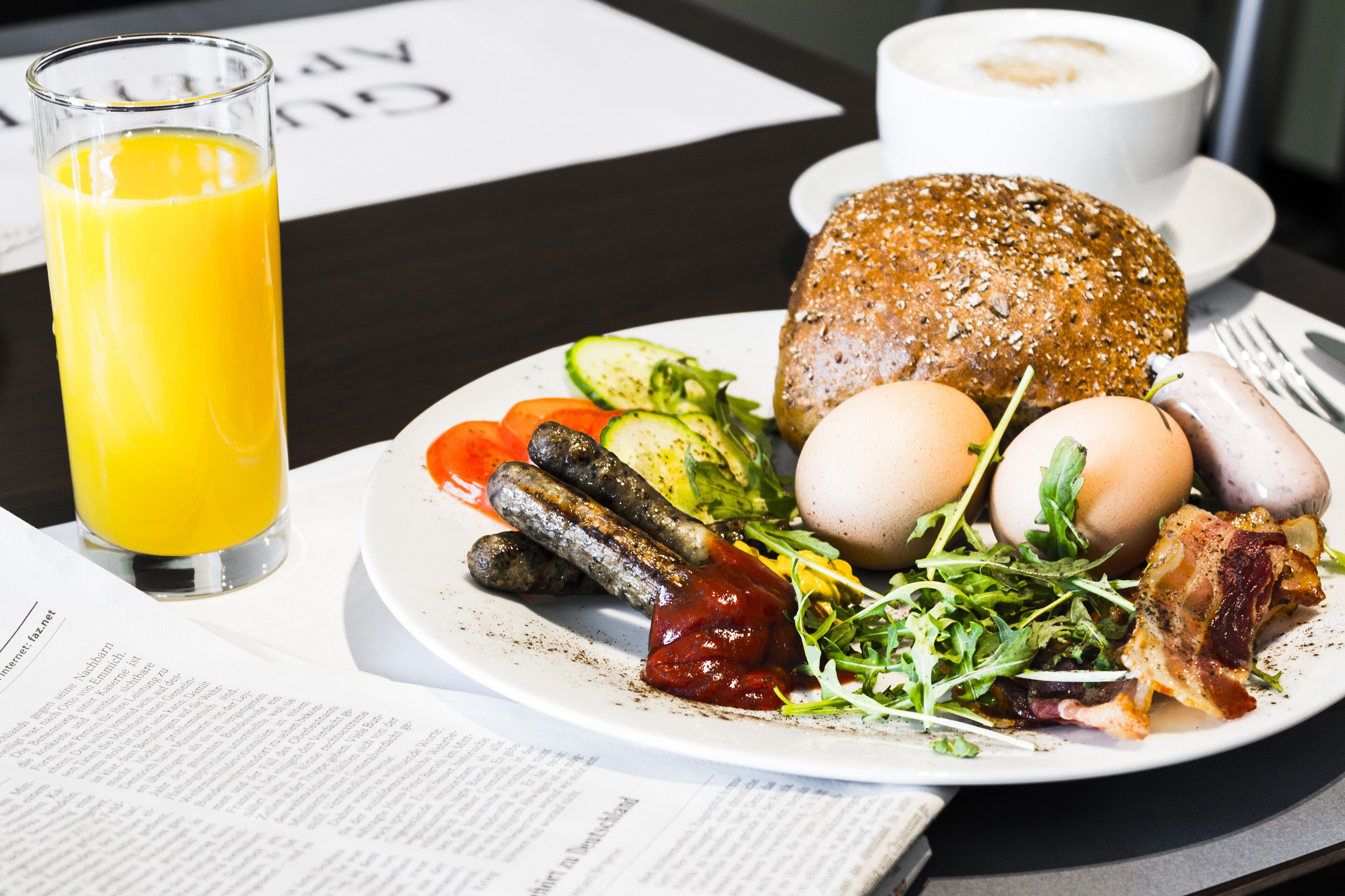 Ein Frühstücksteller mit Eiern, Speck, Würstchen und Gemüse, ein Glas Orangensaft und eine Zeitung auf einem Tisch.