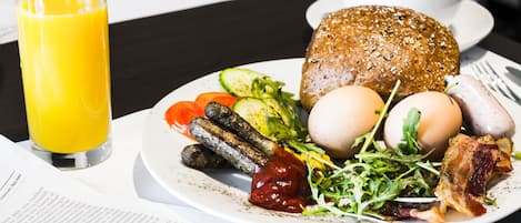 A breakfast plate with eggs, bacon, sausages, and vegetables, a glass of orange juice, and a newspaper on a table.