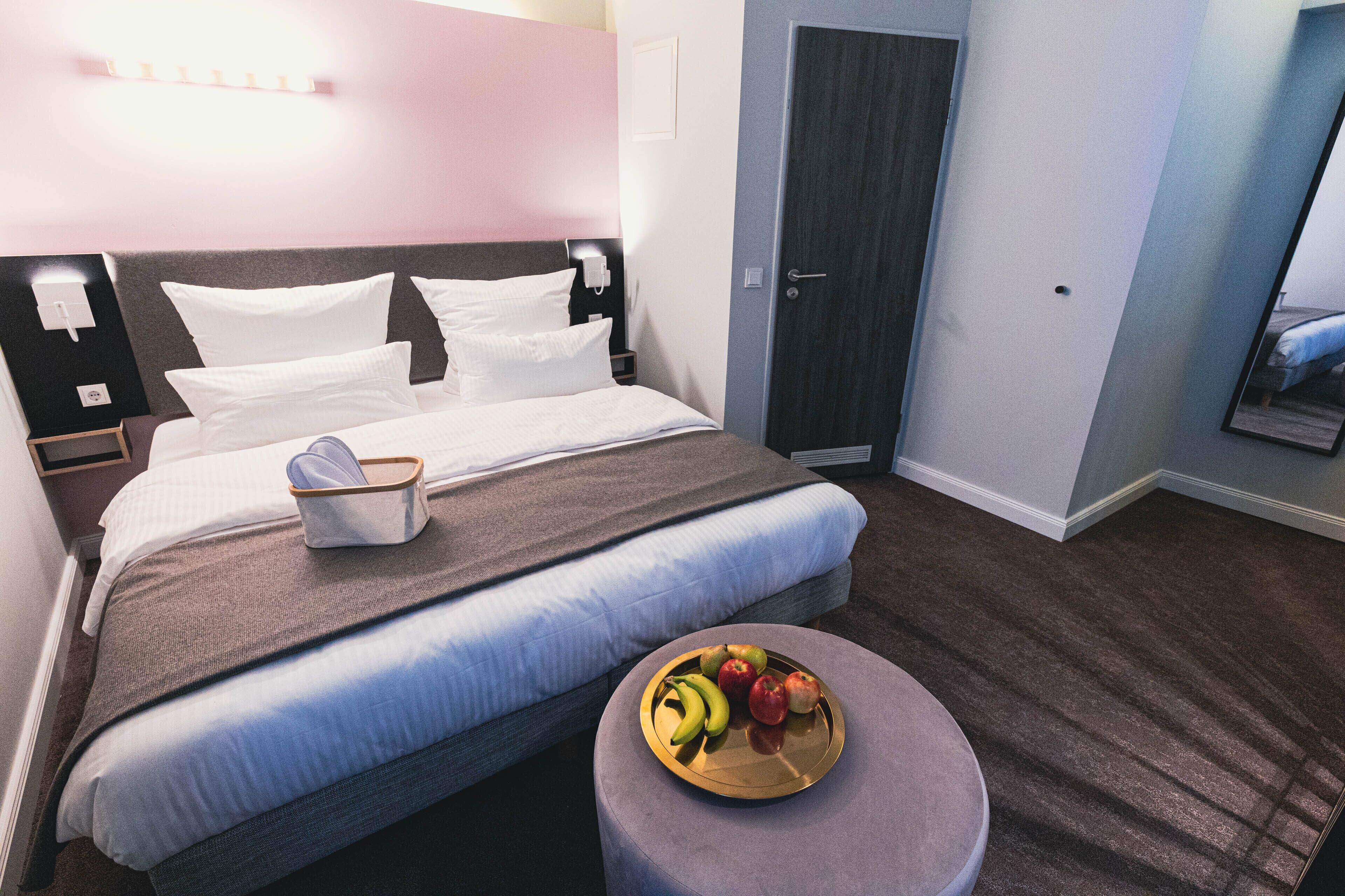 A neatly made bed with white and gray linens, a bedside table with a tray of fruit, and a dark door in a hotel room.