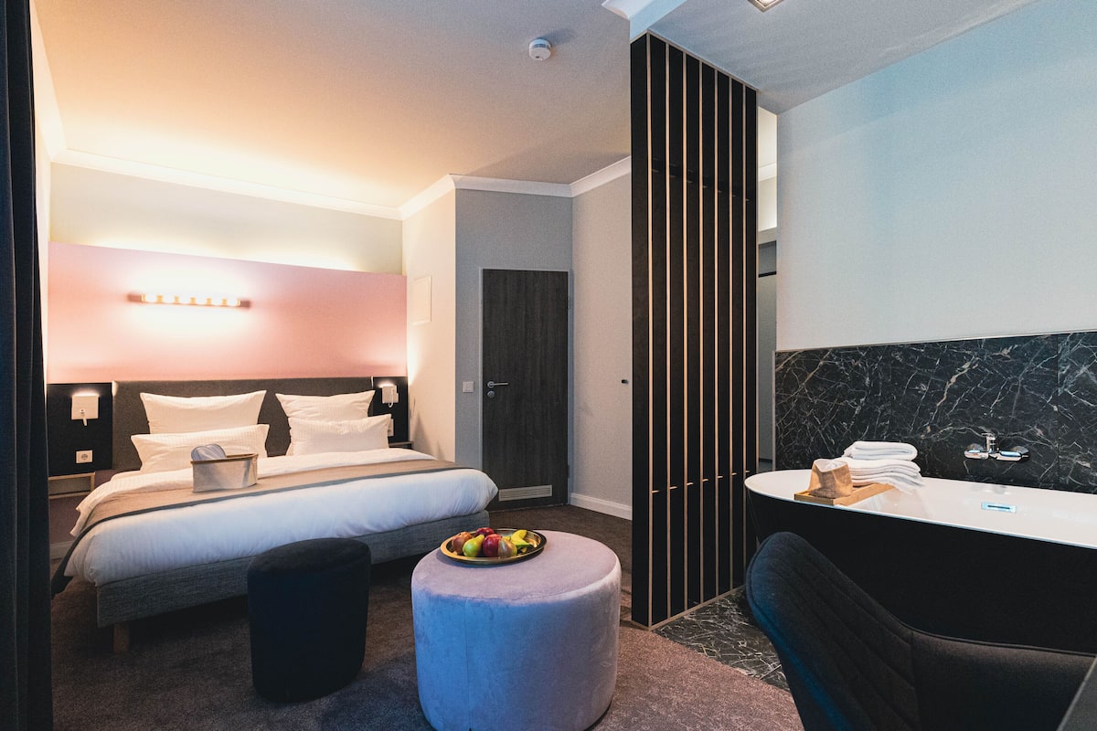 A modern hotel room with a bed, a round ottoman with fruit, a bathroom with a marble wall, and a dark-colored chair.