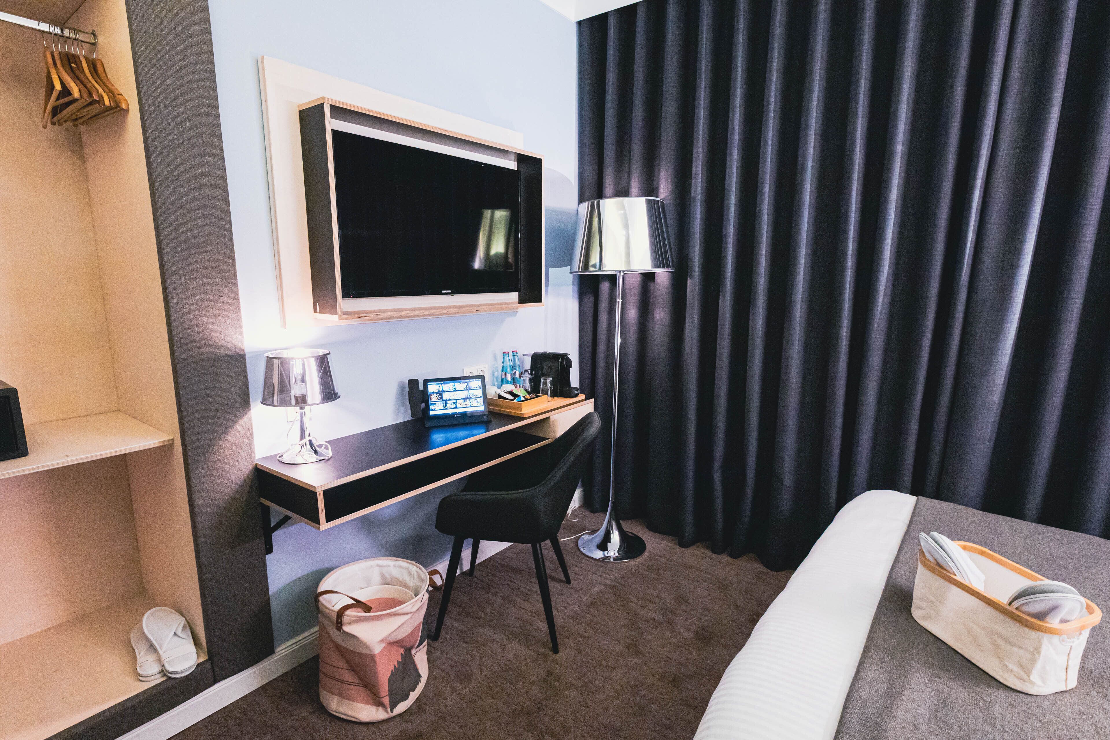 A hotel room with a bed, a desk with a computer, a TV, a lamp, and a window with curtains.