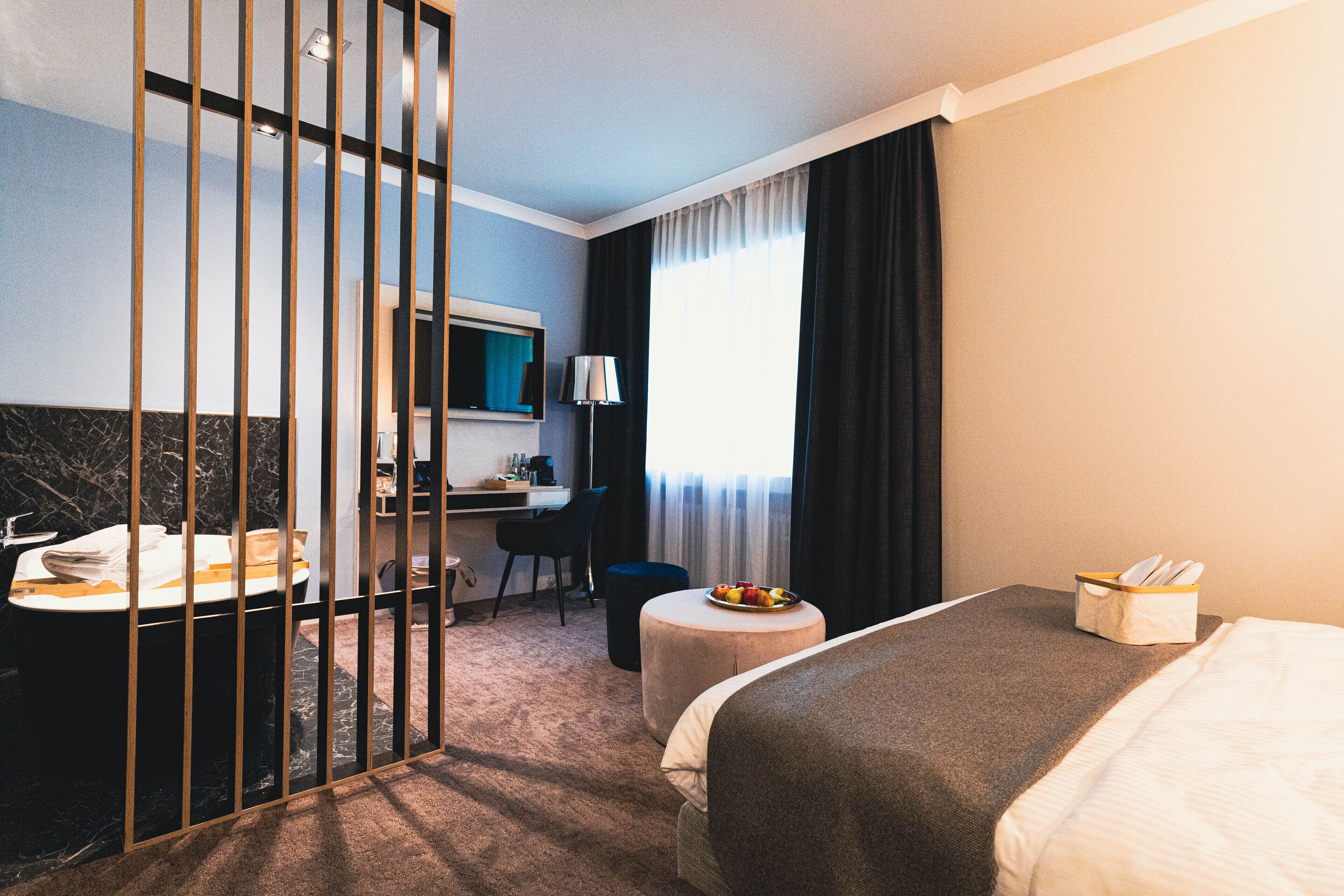 A hotel room with a bed, a desk with a chair, a small round table with a fruit bowl, a bathroom with a sink and mirror, and a window with curtains.