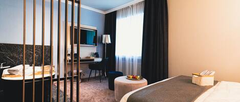 Une chambre d’hôtel comprenant un lit, un bureau avec une chaise, une petite table ronde avec un panier de fruits, une salle de bain avec un lavabo et un miroir, et une fenêtre avec des rideaux.