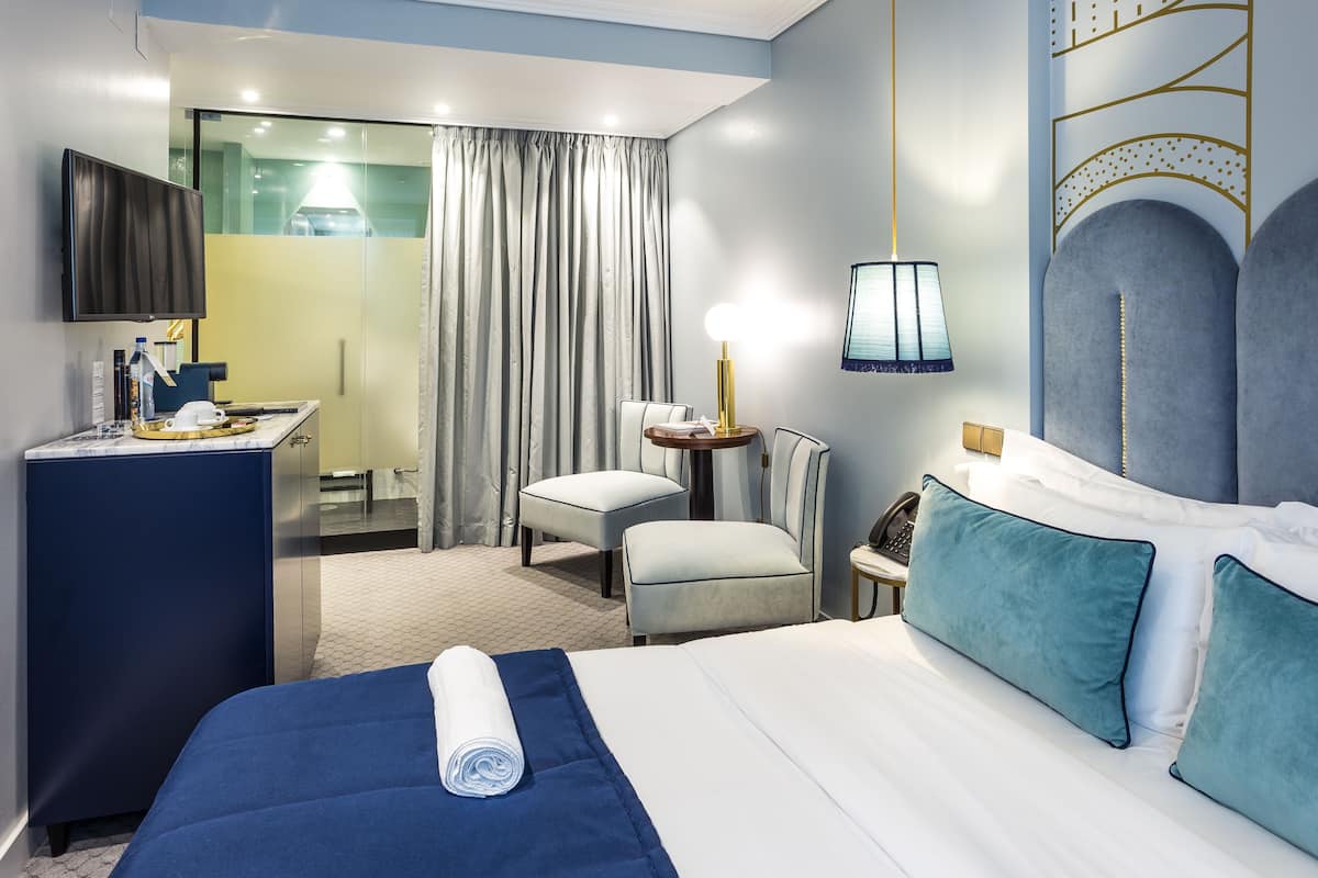 Habitación de hotel con cama, escritorio, silla, televisor y baño visible a través de un separador de vidrio.