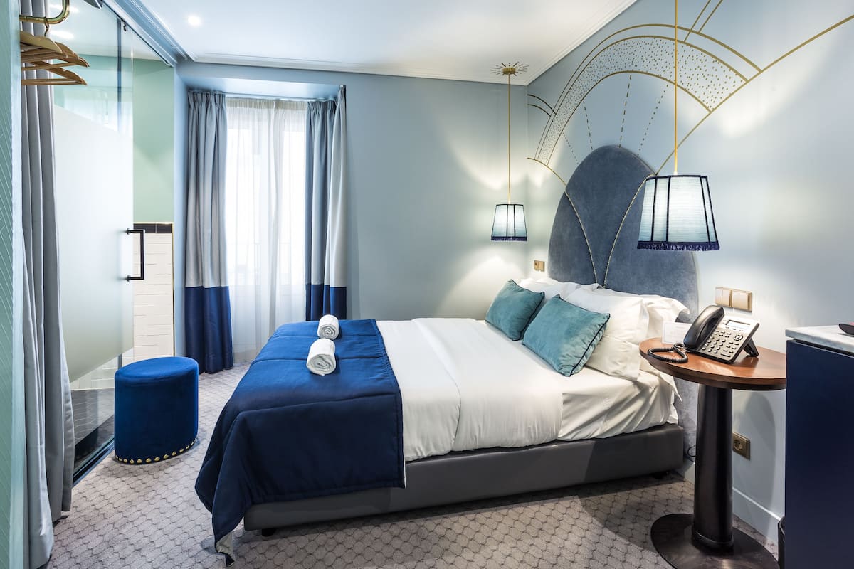 Una habitación de hotel moderna con una cama, un otoman azul redondo, una mesita de noche con un teléfono y una lámpara fijada a la pared.