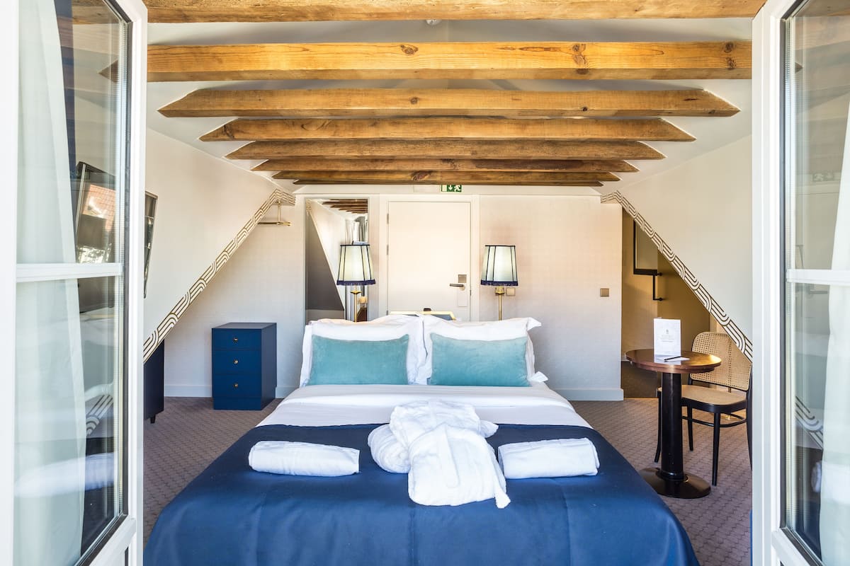 Una habitación de hotel con una cama grande, vigas de madera en el techo, un escritorio pequeño con una silla y una ventana.