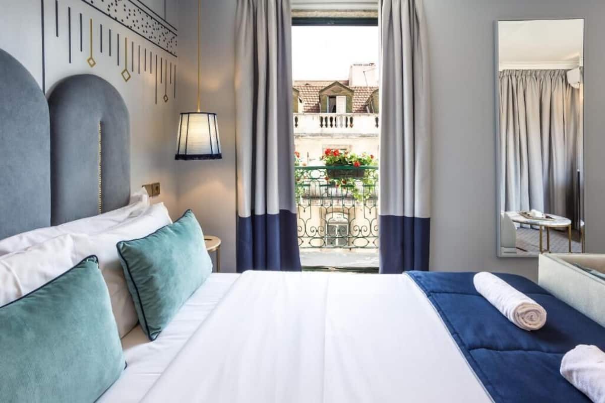Habitación de hotel con una cama grande, una mesita pequeña, un espejo y un balcón con barandilla y algunas plantas.
