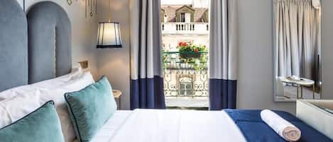 A hotel room with a large bed, a small table, a mirror, and a balcony with a railing and some plants.