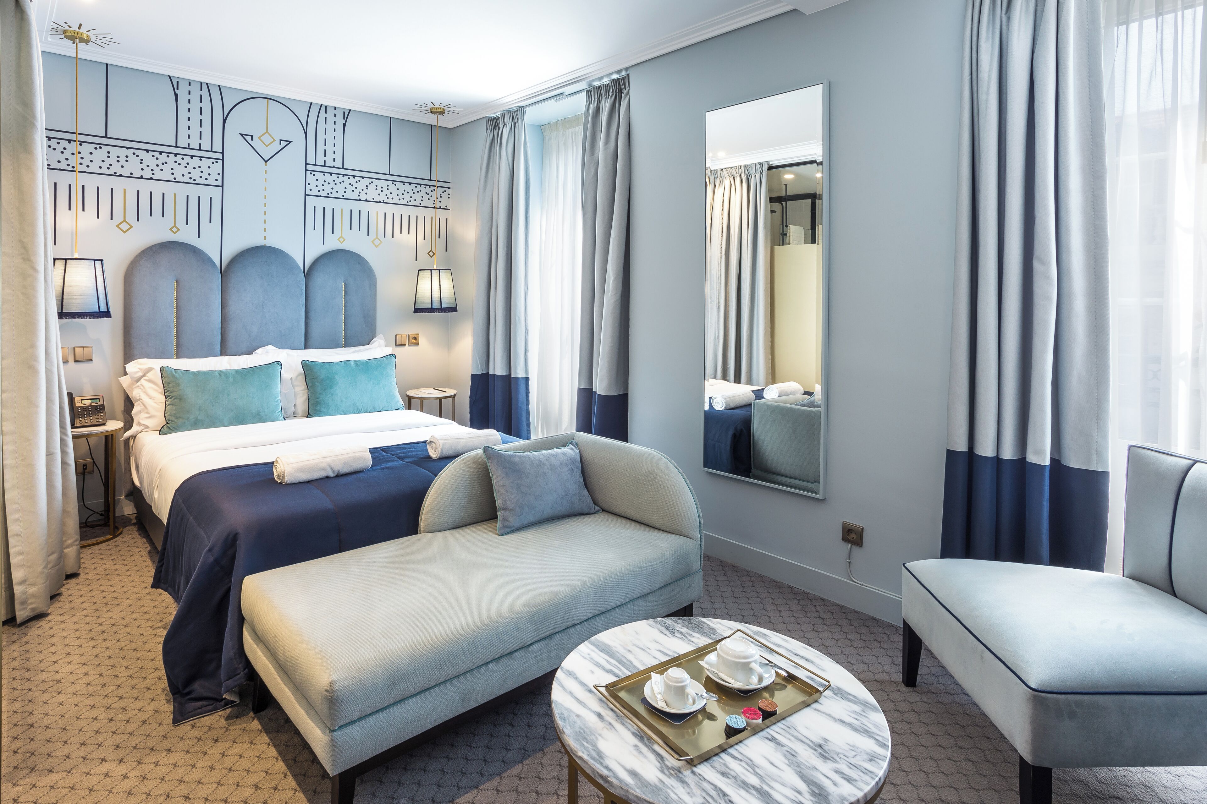 A modern hotel room with a large bed, a sofa, a small table with a tray of items, and a large mirror.