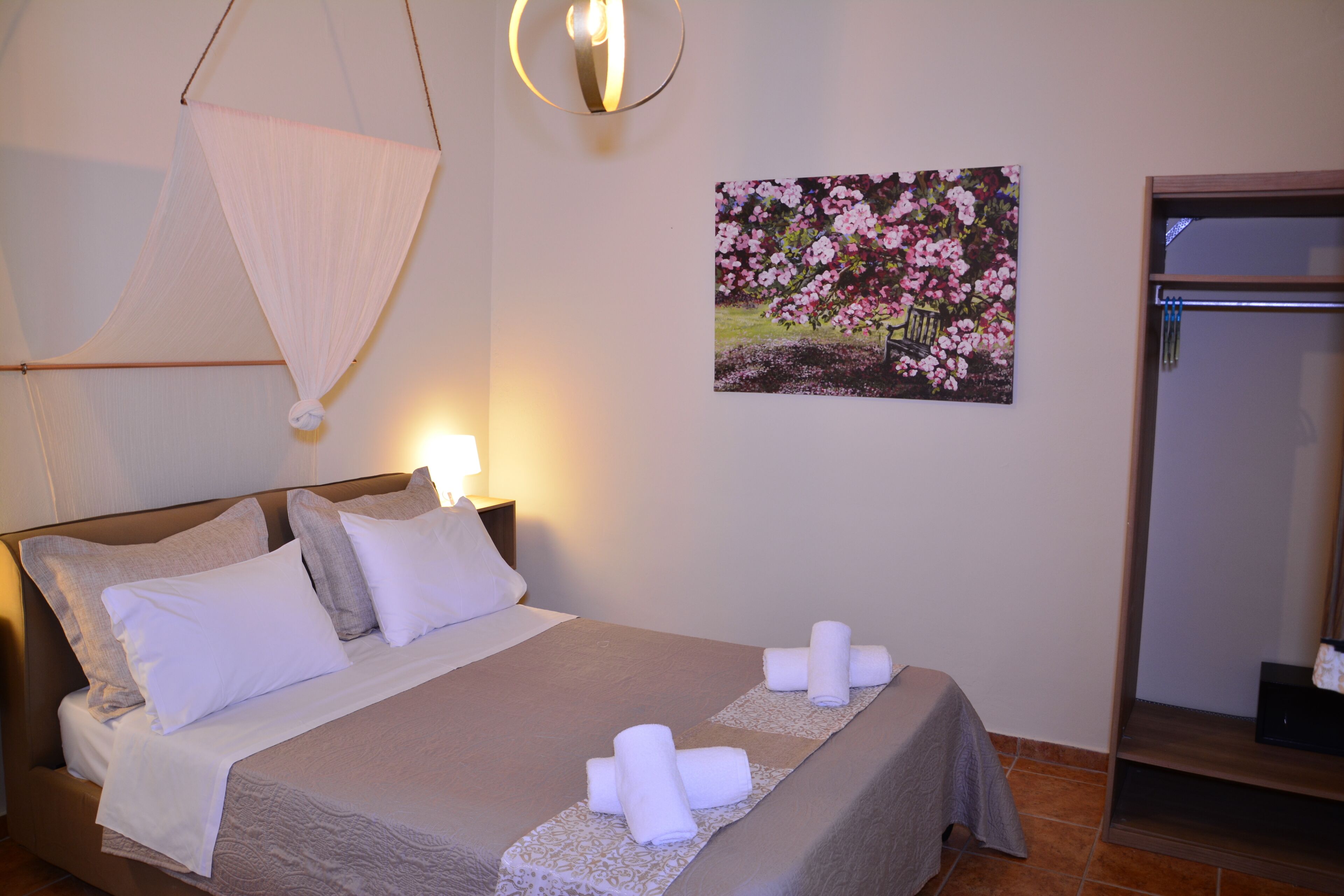 Cama bem arrumada com travesseiros, luminária fixada na parede, pintura de flores e porta com porta-toalhas.