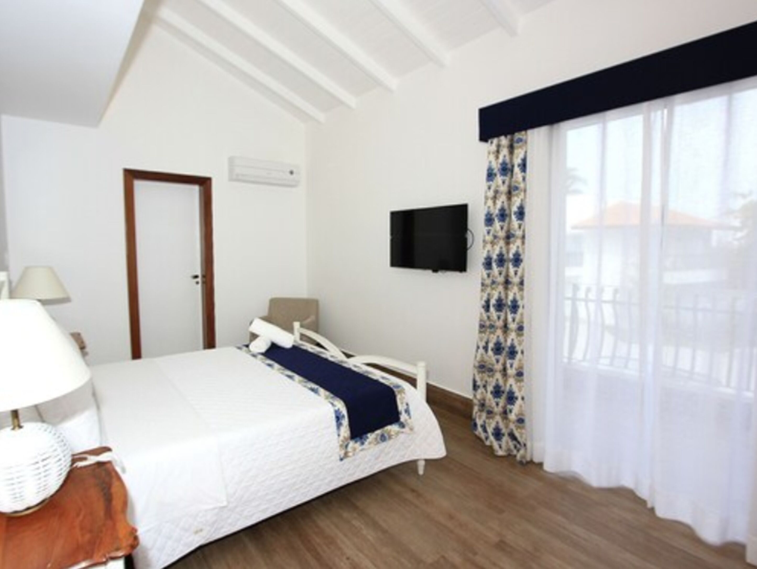 A bedroom with a bed, a TV mounted on the wall, a door, and a window with curtains.