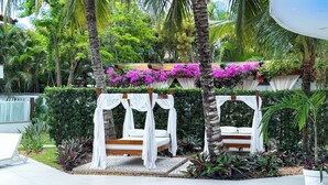 Un área junto a la piscina con dos camas colgantes de madera cubiertas de tela blanca, rodeada de exuberante follaje y plantas con flores.