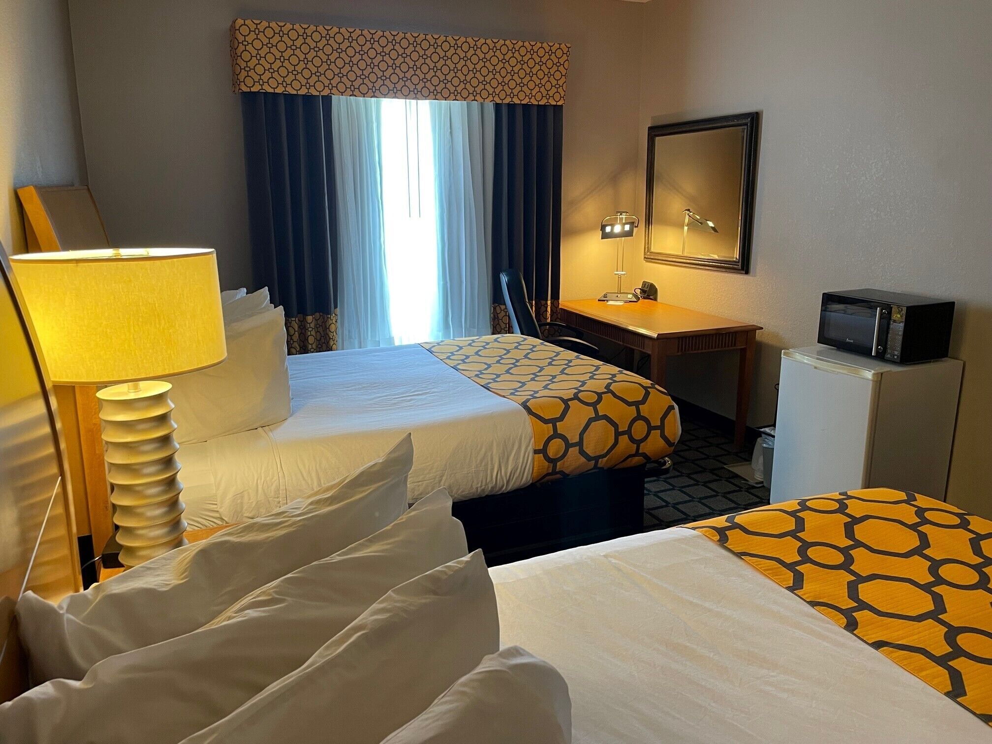 A hotel room with two beds, a desk, a chair, a lamp, a mirror, a microwave, and a window with curtains.