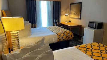 A hotel room with two beds, a desk, a chair, a lamp, a mirror, a microwave, and a window with curtains.