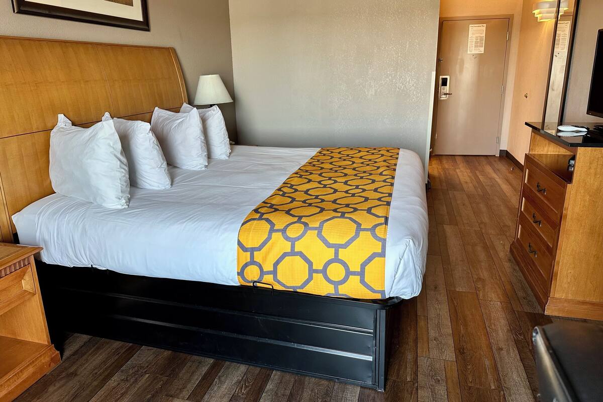 Habitación de hotel con cama, cabecero de madera, mesita de noche, lámpara y cómoda con cajones.