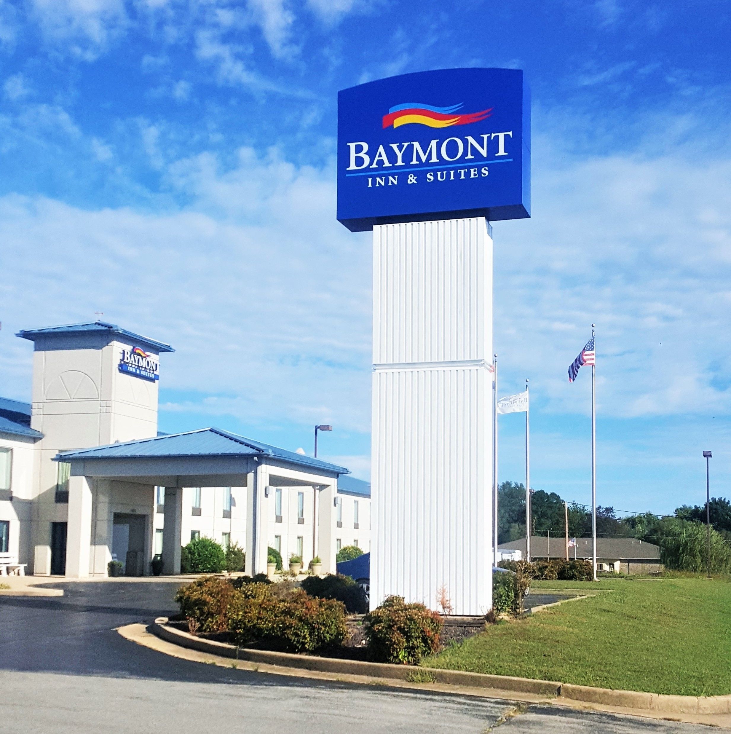 A Baymont Inn & Suites sign with a clear view of the building's entrance, a flagpole with the American flag, and a well-maintained lawn.