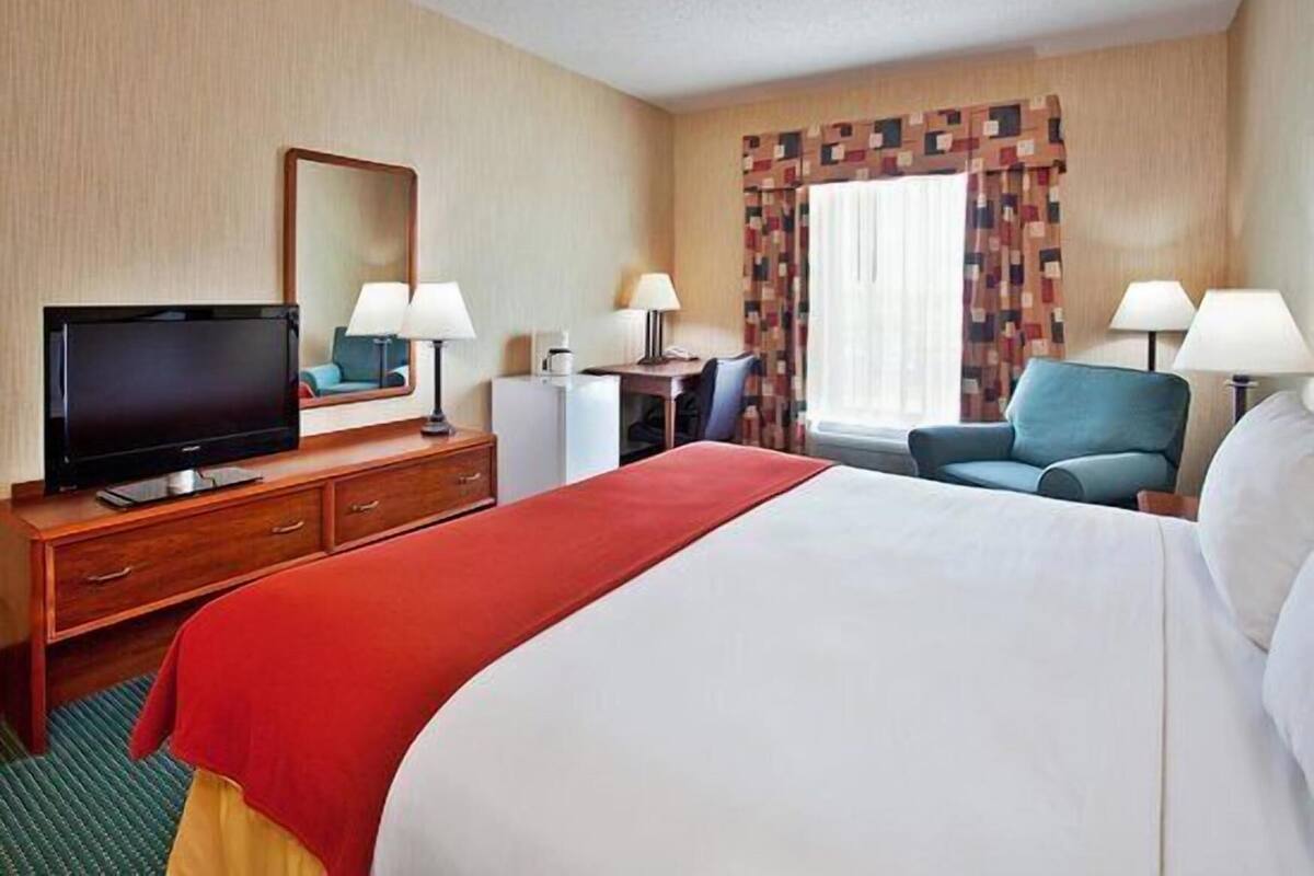 Habitación de hotel con cama, escritorio con televisor, dos sillas, lámpara de escritorio y ventana con cortinas estampadas.