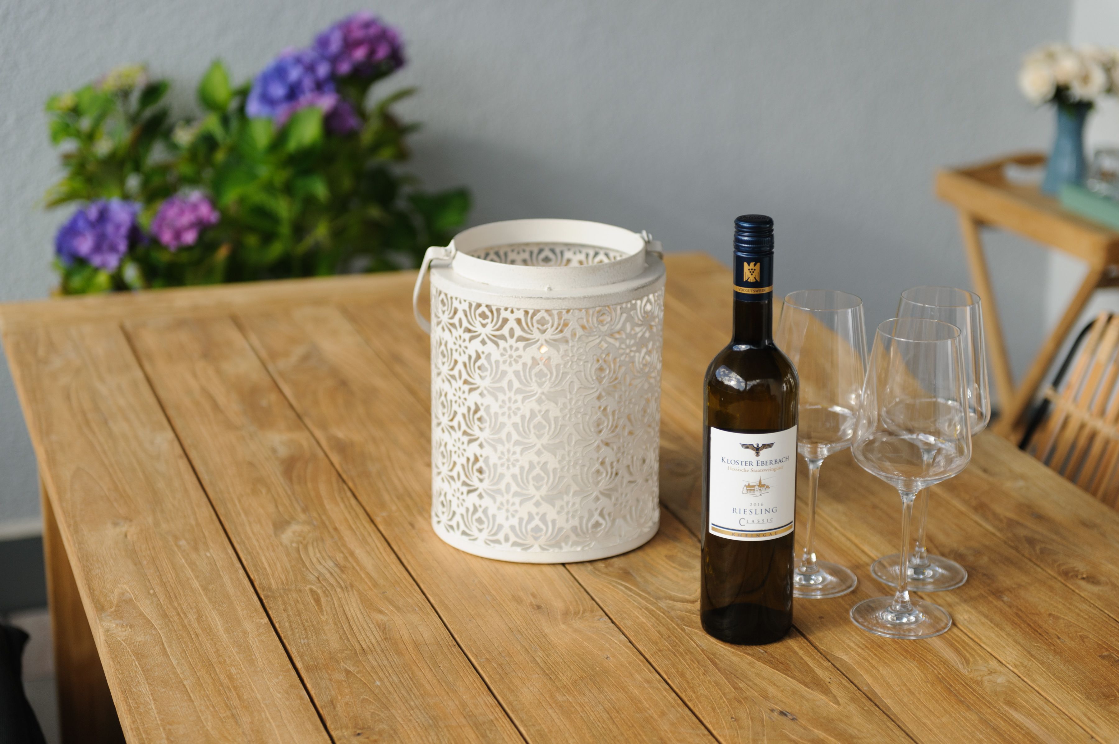 Een houten tafel met een fles wijn en glazen, een witte decoratieve vaas en paarse bloemen op de achtergrond.