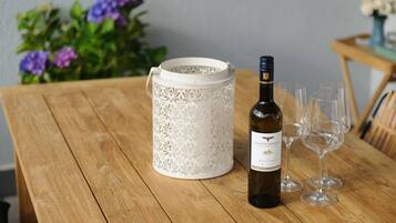 Een houten tafel met een fles wijn en glazen, een witte decoratieve vaas en paarse bloemen op de achtergrond.