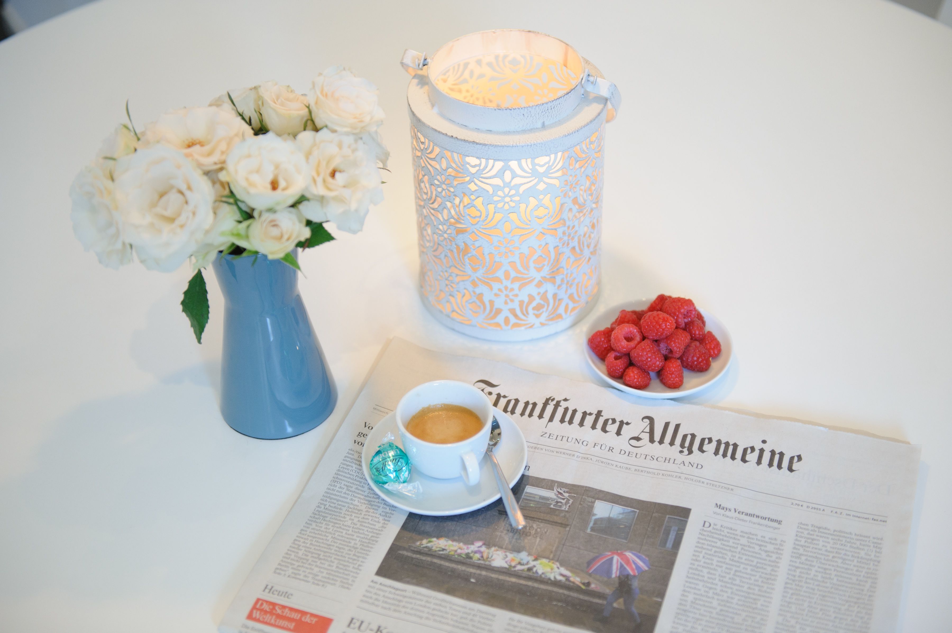 A table with a white vase of white roses, a white candle, a bowl of red raspberries, a cup of coffee, and a newspaper.