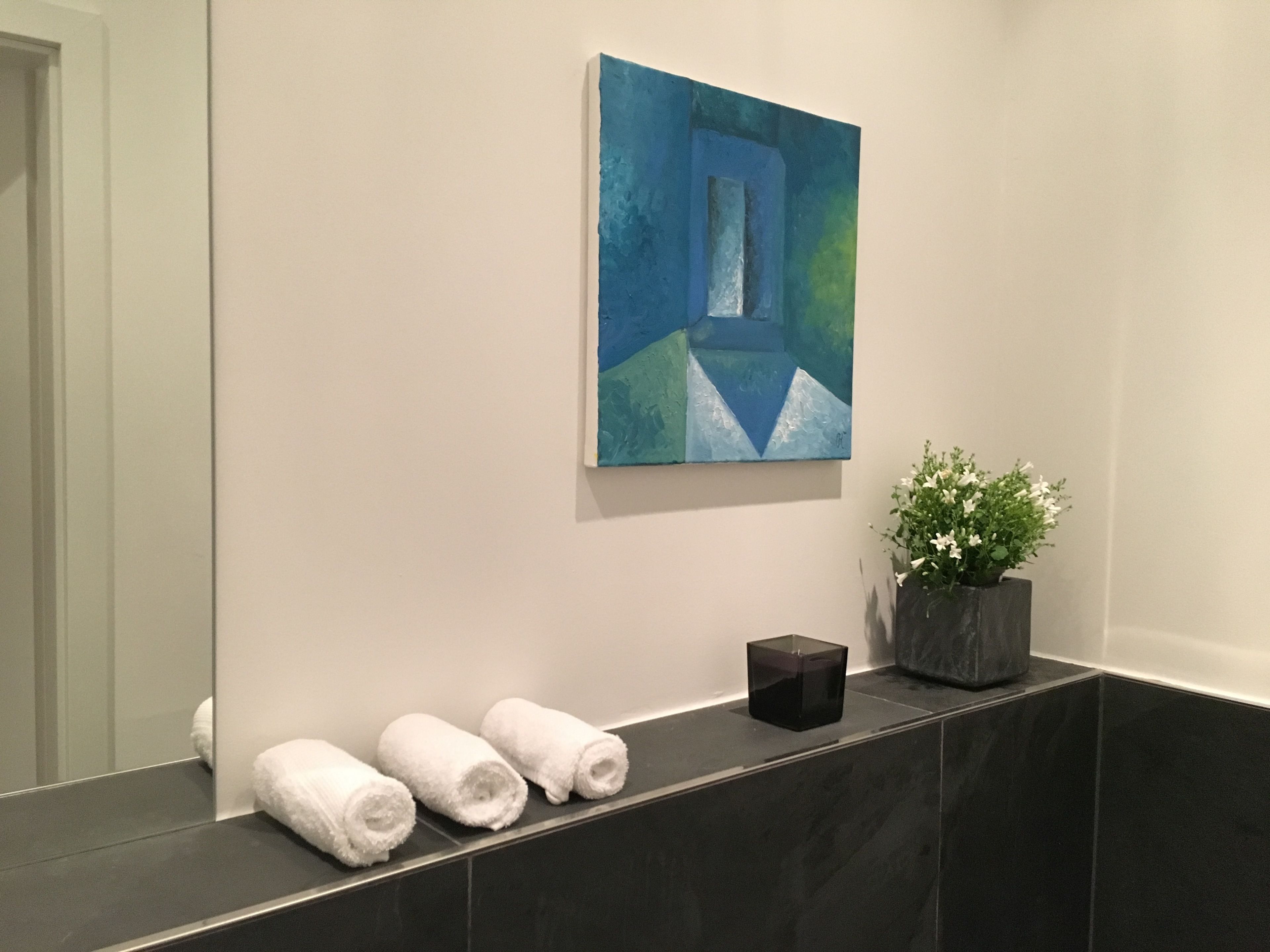Een badkamer met een donkere marmeren aanstalt, drie opgerolde handdoeken, een potplant en een schilderij aan de muur.