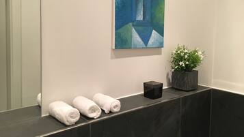 Een badkamer met een donkere marmeren aanstalt, drie opgerolde handdoeken, een potplant en een schilderij aan de muur.
