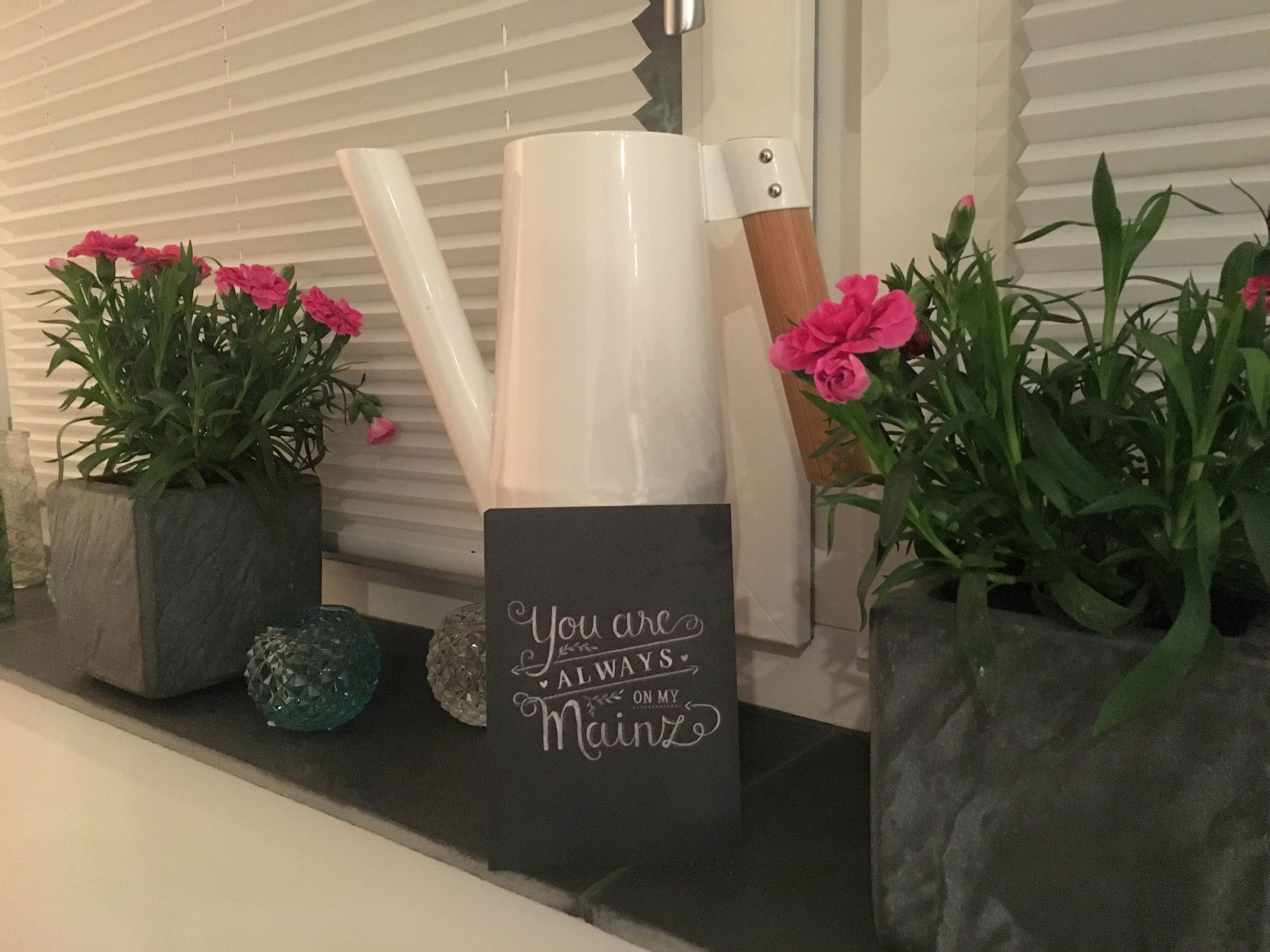 Ein Regal mit Dekorationsobjekten, darunter ein schwarzes Schild mit weißer Schrift, rosafarbene Blumen und eine weiße Vase.