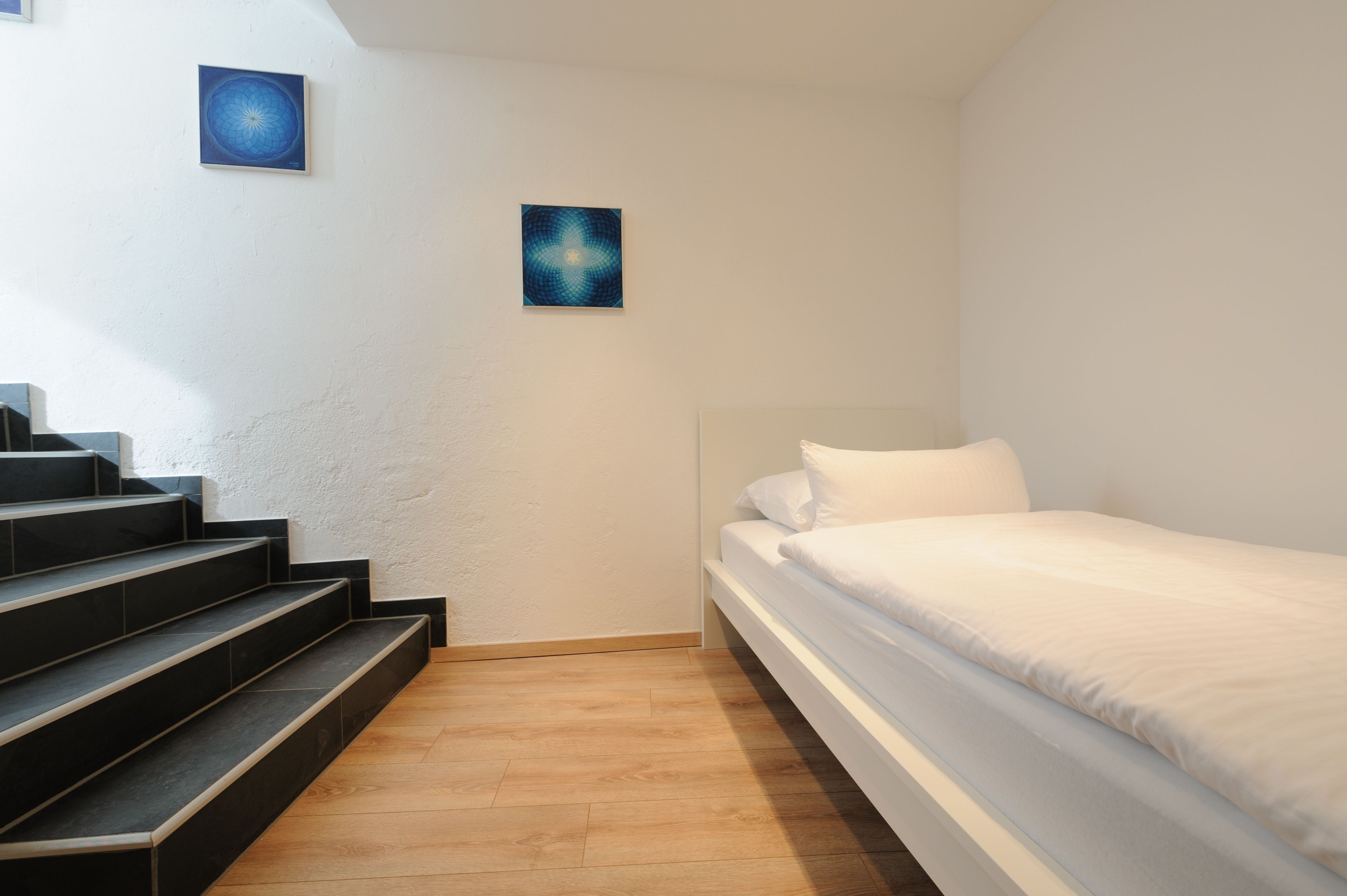 A bedroom with a bed, white walls, a wooden floor, and a set of stairs leading to a balcony.