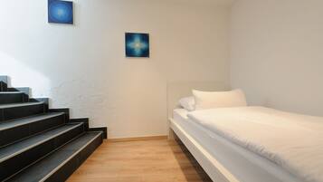A bedroom with a bed, white walls, a wooden floor, and a set of stairs leading to a balcony.