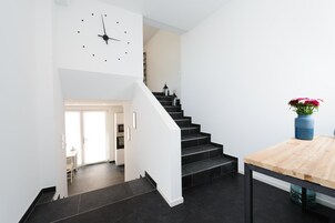 Una escalera moderna con peldaños negros y paredes blancas, que conduce a una habitación con una mesa de madera y un florero con flores.