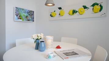 A round dining table with a white tablecloth, a vase with white flowers, a blue vase, a white candle holder, a plate of red berries, a cup with a teapot, and a newspaper.