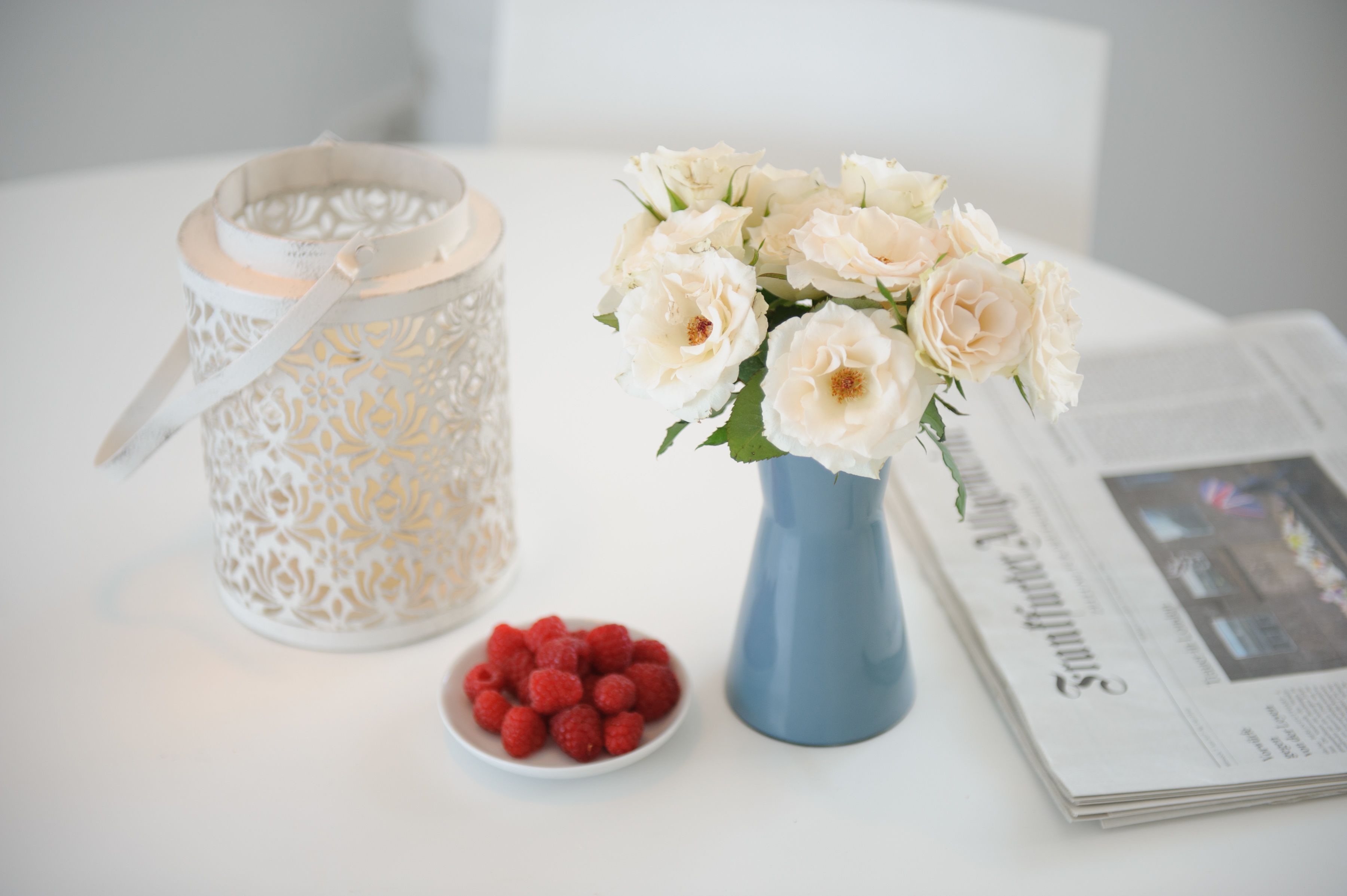 Ein weißer Tisch mit einem weißen Kerzenständer, einer blauen Vase mit weißen Blumen, einer Schüssel voller roter Beeren und einer Zeitung.