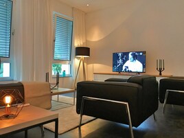 Una sala de estar moderna con un televisor de pantalla plana, una lámpara de pie y un comedor con mesa y sillas.