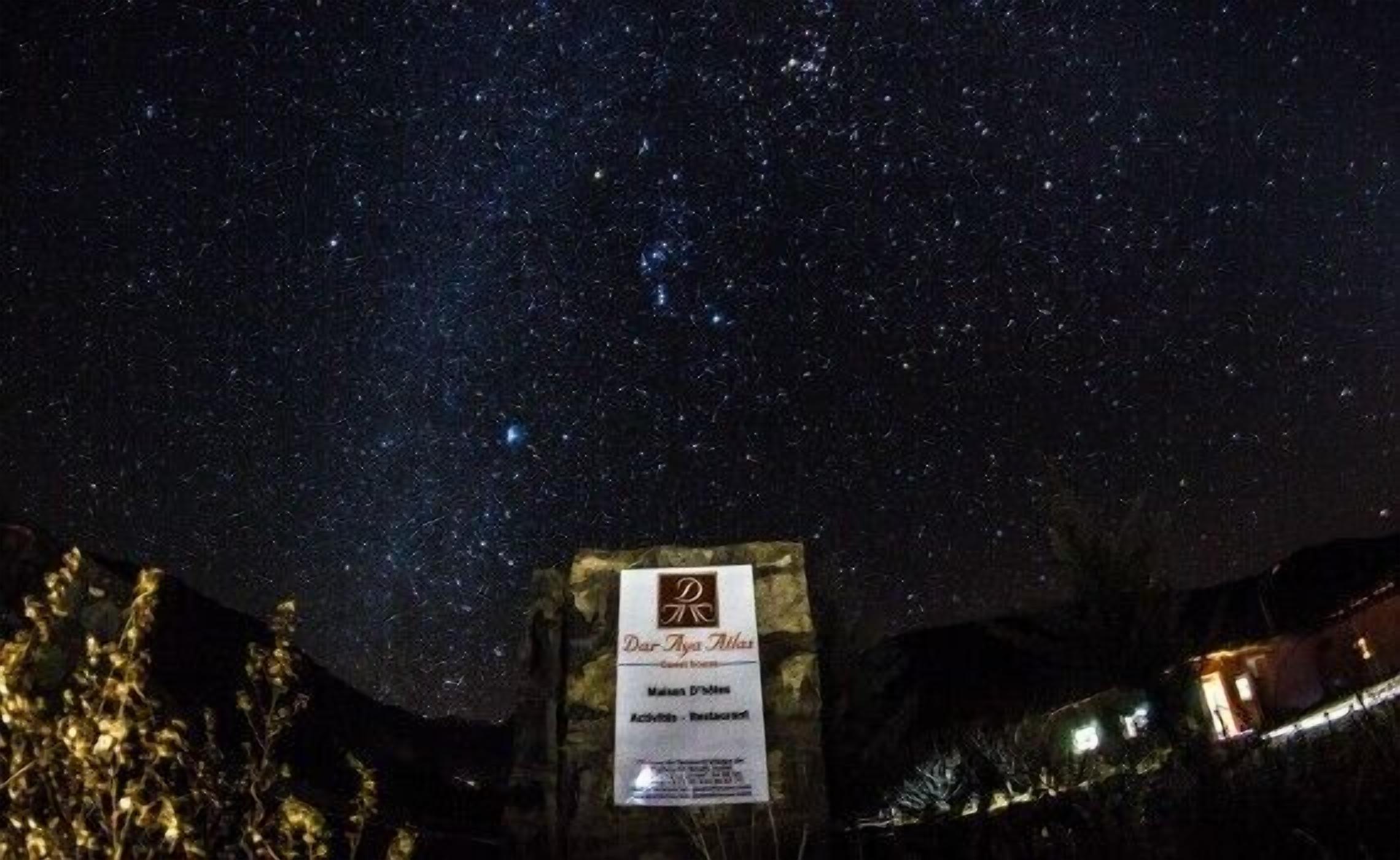 A night sky filled with stars above a sign for 'Der Aya Atlas' with a view of a mountainous landscape.