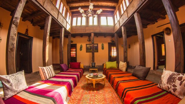 A traditional inn with wooden beams, a central round table, and colorful striped bedding. - Dar Aya Atlas (Ait Bou Oulli)