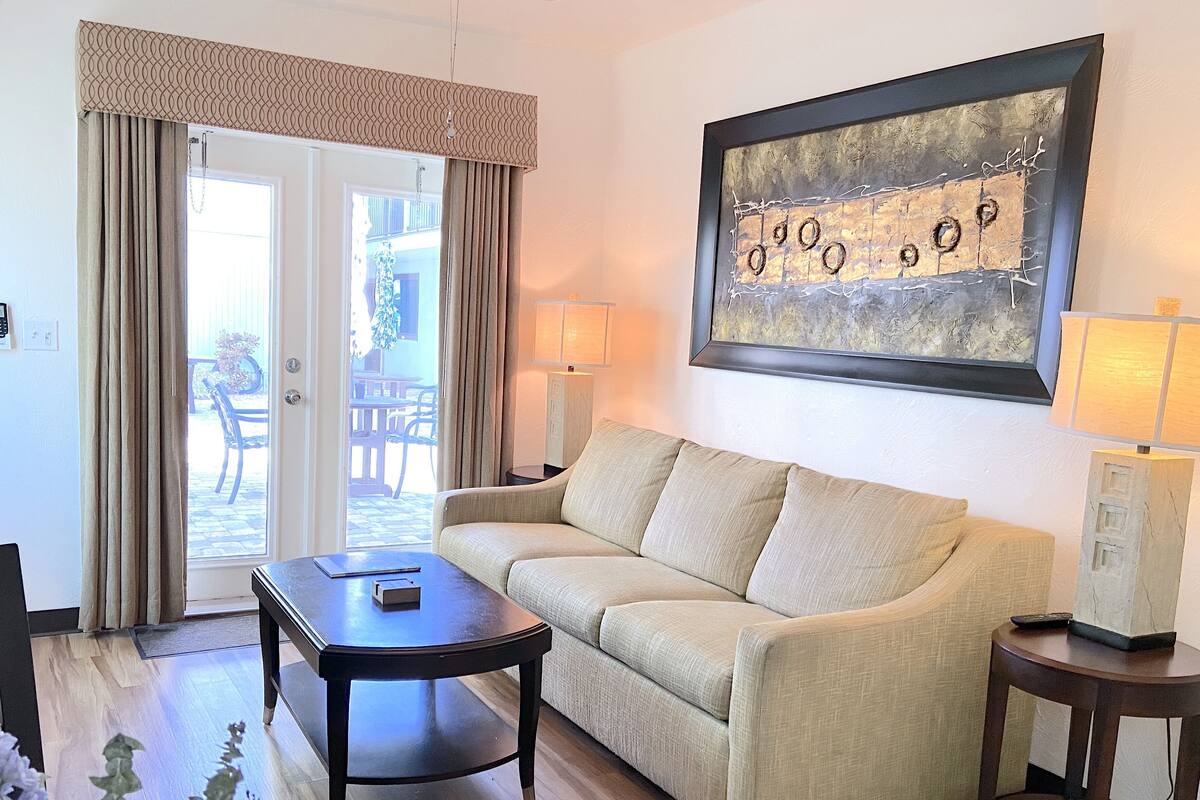 A living room with a sofa, a coffee table, and two lamps. There is a door leading to an outdoor area with a table and chairs.