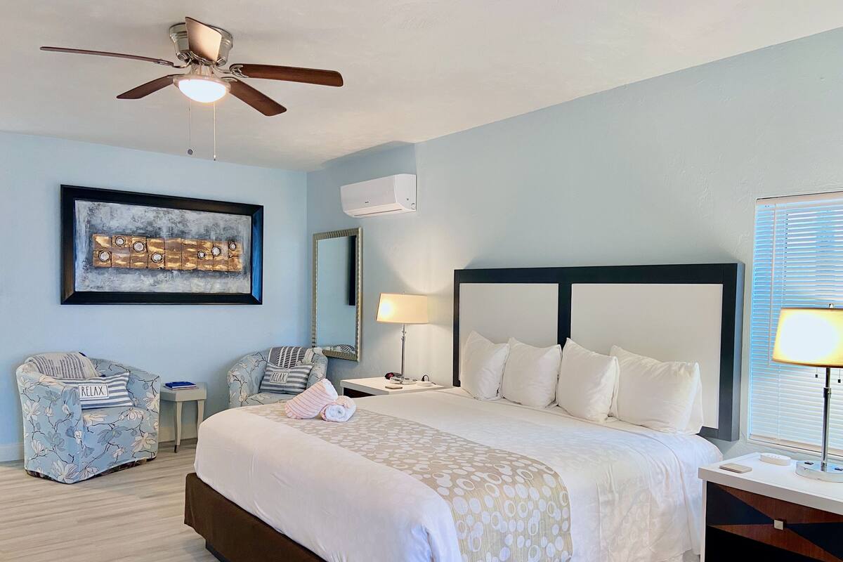 A hotel room with a bed, two armchairs, a ceiling fan, a wall-mounted air conditioner, a framed artwork, a bedside lamp, and a window with blinds.
