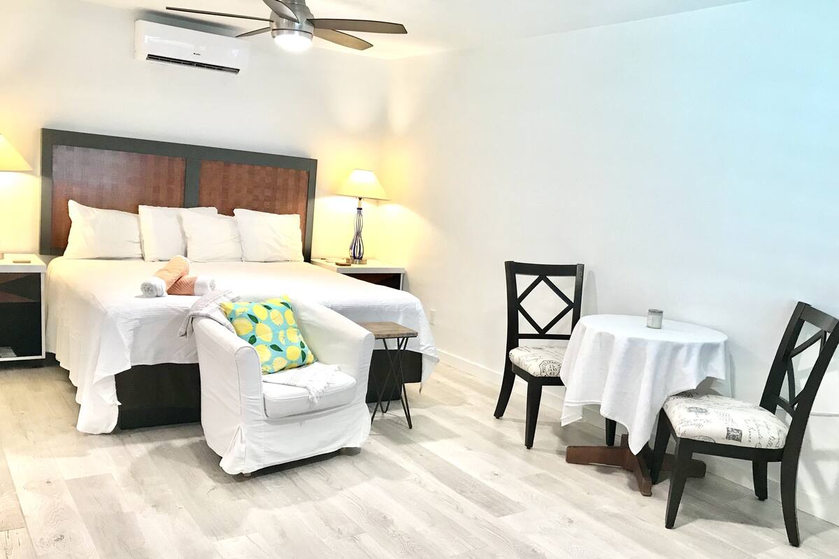 A hotel room with a bed, a white armchair, a wooden chair, a small table, and a ceiling fan.