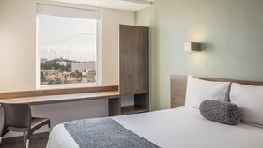 A hotel room with a bed, a desk, a chair, a window with a city view, and a wall-mounted light. - One Puebla Serdan (Puebla)