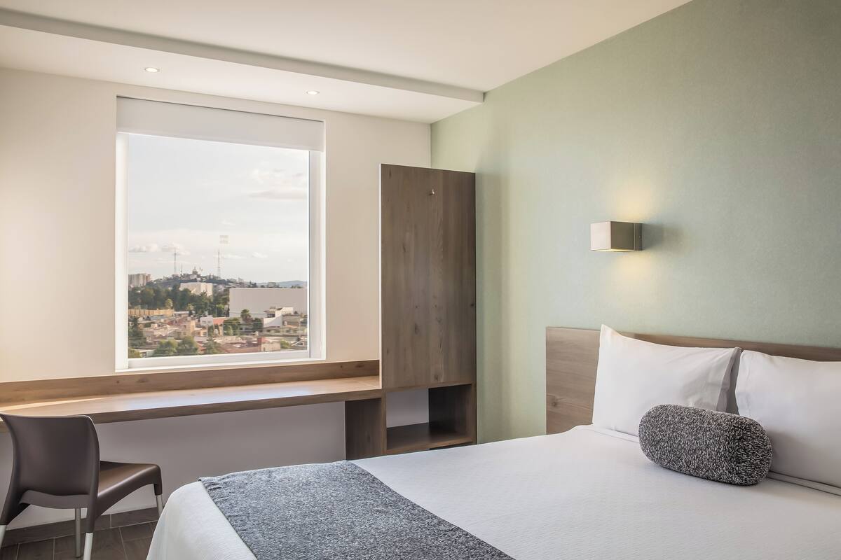 A hotel room with a bed, a desk, a chair, a window with a city view, and a wall-mounted light.