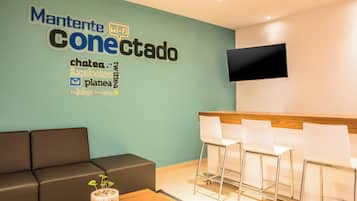 Un área de recepción con una pared color turquesa, un sofá negro, una mesa de madera con una planta y una zona de bar con sillas blancas.