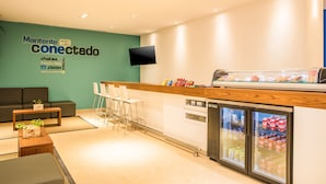 A modern reception area with a green wall, a wooden counter, seating, and a refrigerator stocked with beverages. - One Puebla Serdan (Puebla)