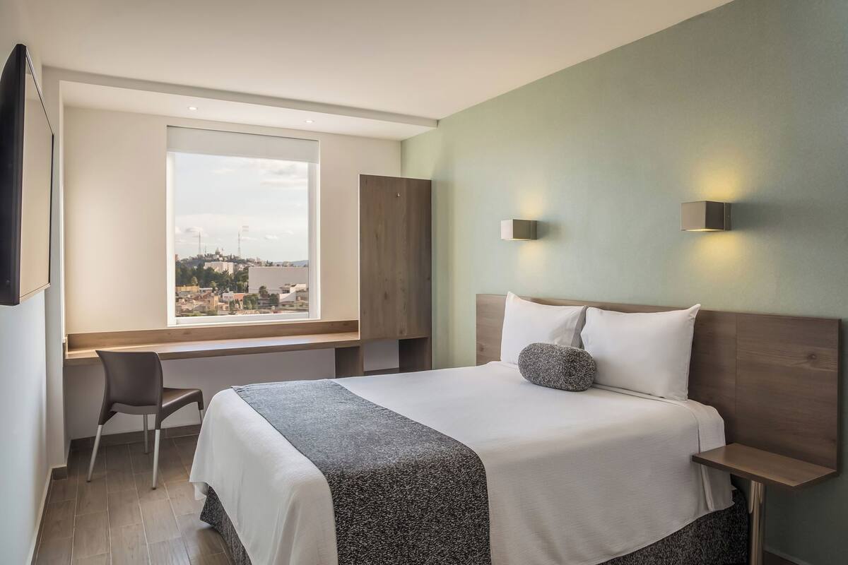 A hotel room with a bed, a desk, a chair, a window with a city view, and two wall-mounted lights.