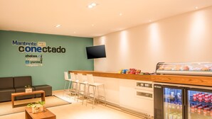 A modern reception area with a seating area, a counter with snacks, a TV mounted on the wall, and a refrigerator stocked with beverages. - One Puebla Serdan (Puebla)