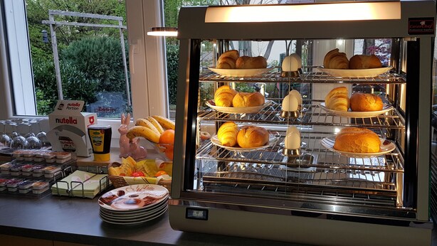A bakery display case with pastries and bread, a Nutella dispenser, and a window view of greenery. - H41 Inn Freiburg (Gundelfingen)
