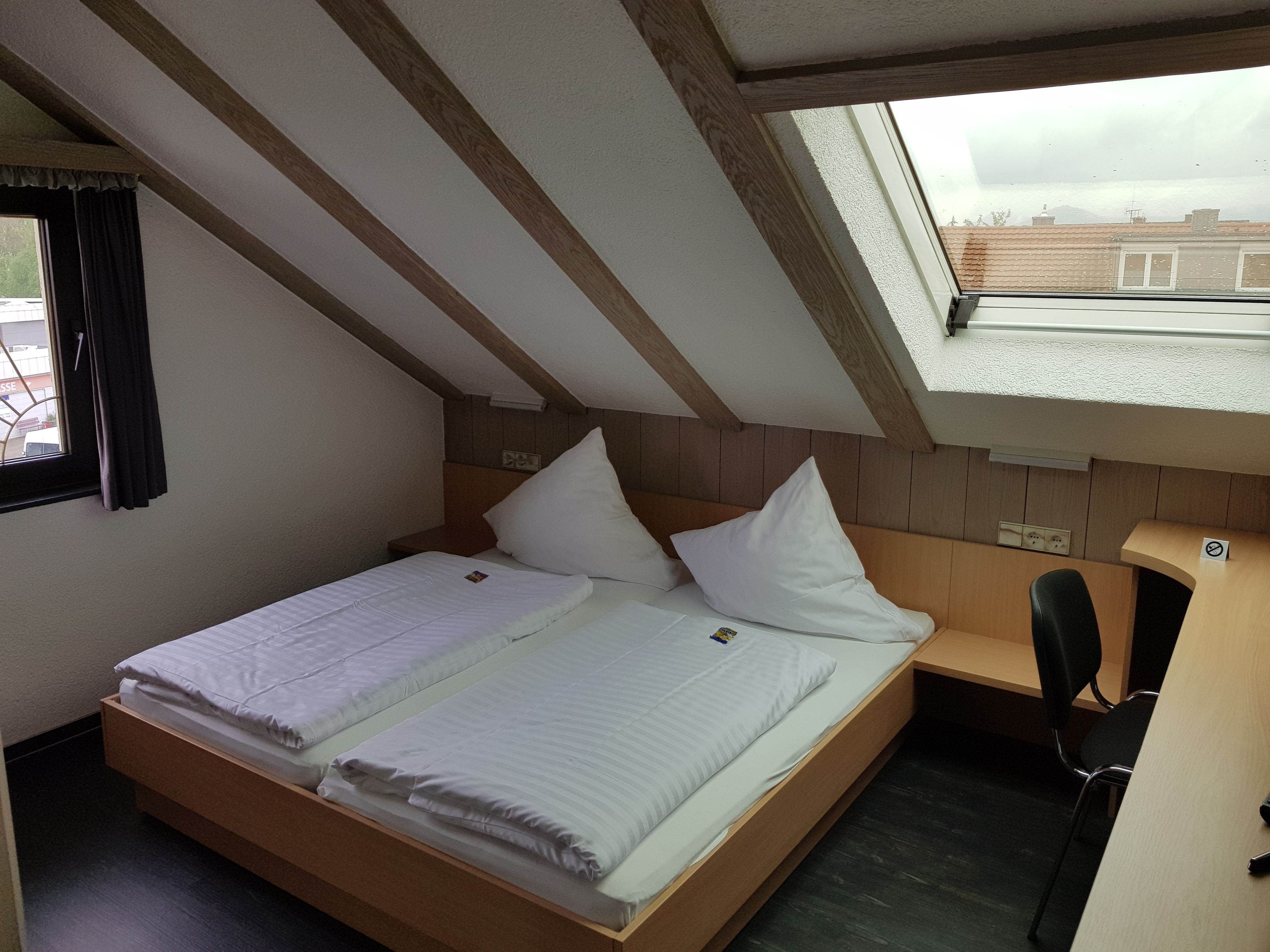 Ein kleines Zimmer mit einem Einzelbett, einer schrägen Decke und einem Fenster mit Blick auf ein Gebäude.