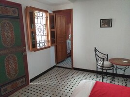 Um quarto com uma cama, uma mesinha com uma cadeira, uma porta de madeira com painel de vidro decorativo e piso de azulejos.