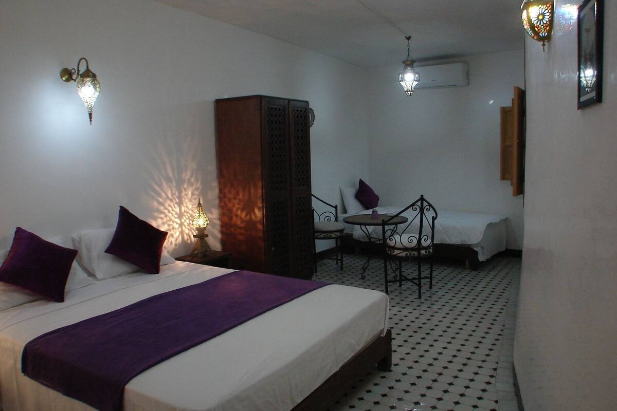 Una camera d'albergo con un letto, un tavolino con una sedia, un armadietto e due luci a parete.