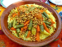 Uma grande tigela redonda e marrom cheia de cuscuz, grão de bico, cenouras, abobrinha e outros vegetais.