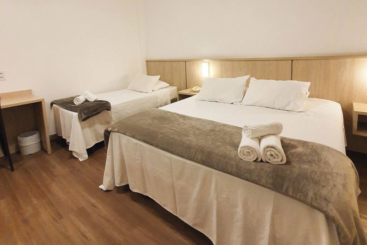 Habitación de hotel con dos camas, cabecera de madera, mesita de noche con lámpara y un cubo de basura pequeño.
